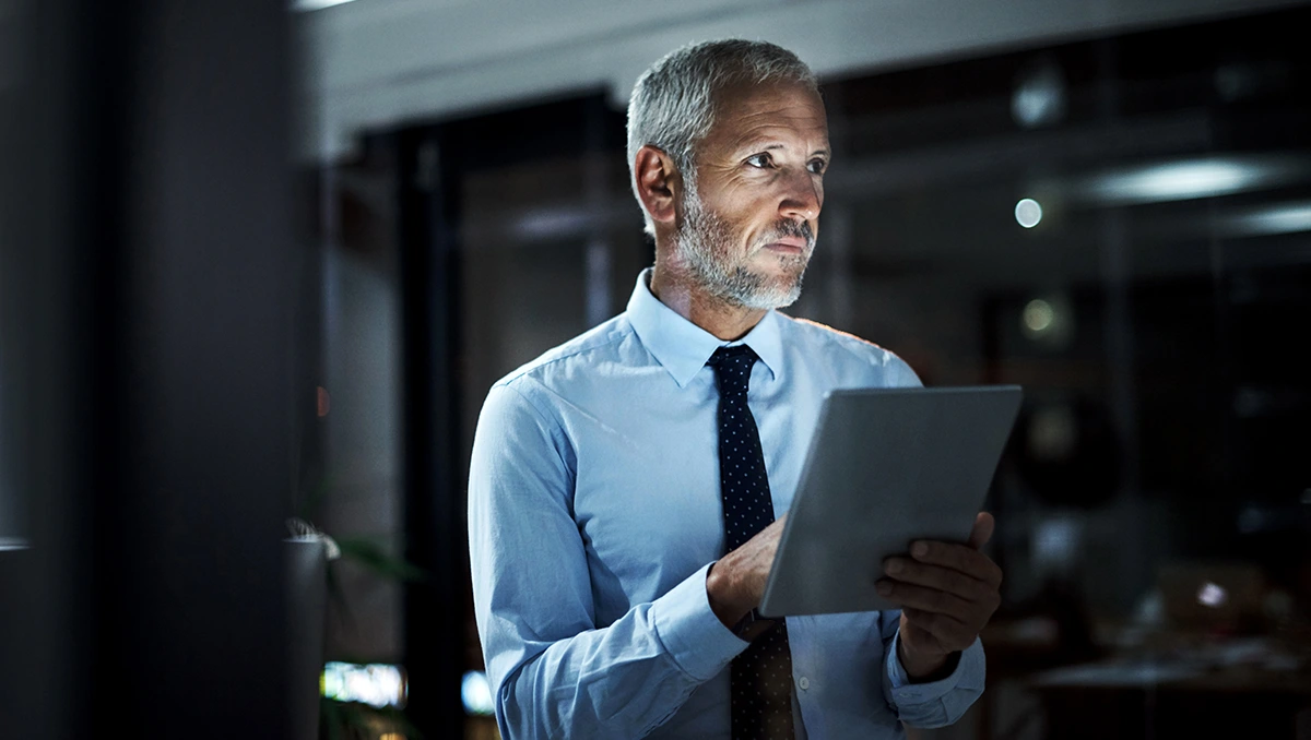 Middle-aged business professional standing in a dark office space holding a tablet device